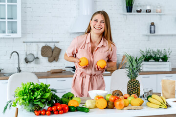 A woman is smiling and holding two oranges in her hands. She is standing in front of a table full of fruits and vegetables