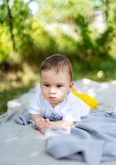 A baby is laying on a blanket in a park. The baby is wearing a yellow shirt and is looking at the camera.