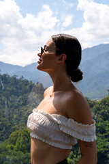 Side profile of a woman in sunglasses and a white off-shoulder top, enjoying the scenic view of lush green mountains under a clear sky. The scene is peaceful and natural.