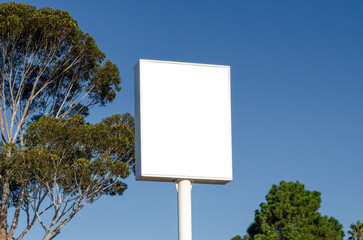Blank white mockup background texture of a tall vertical rectangle retail shop sign. Empty store ad template against sky and trees in natural sunlight.