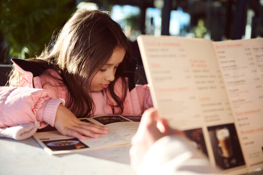 Little girl in a pink jacket choosing a meal from the menu at a cafe