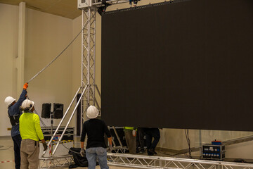 Three technical workers dressed in safety clothing work assembling an LED screen in an auditorium.