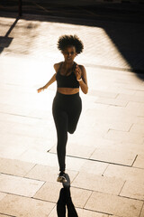 Woman running with determination on a sunlit urban street. Woman with an athletic posture jogging...