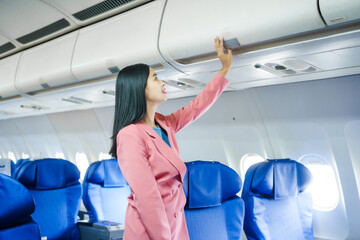 A young Asian female airplane passenger sits by the window during the flight, holding her boarding pass, with her carry-on luggage stored overhead, wearing her seatbelt, and enjoying the journey.