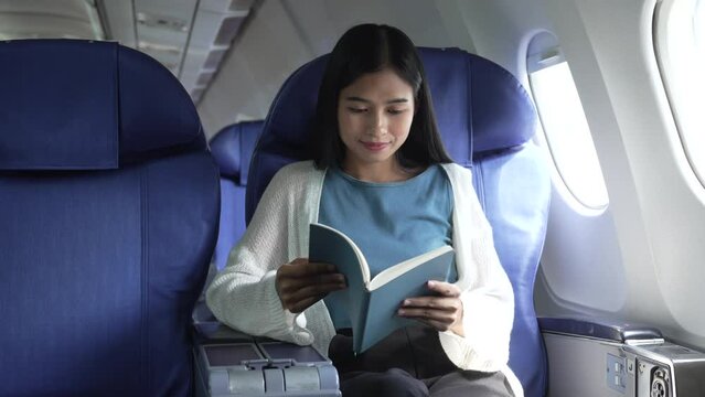 A young Asian female airplane passenger sits by the window during the flight, holding her boarding pass, with her carry-on luggage stored overhead, wearing her seatbelt, and enjoying the journey.