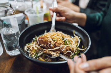 A bowl of noodles with ingredients such as chicken thighs, bean sprouts, and chopped onions in a black bowl. One hand holds a fork and spoon.