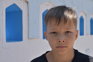 A serious fair-haired boy against the background of a white-blue fence with geometric shapes.