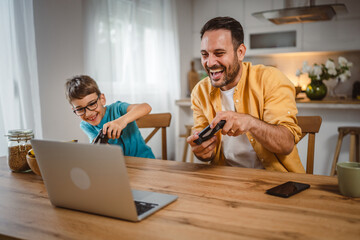 Father and son play video games on laptop with joystick at home
