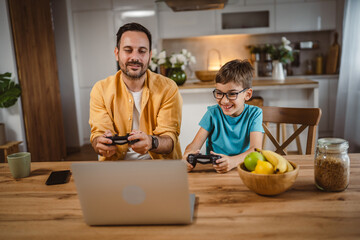 Father and son play video games on laptop with joystick at home