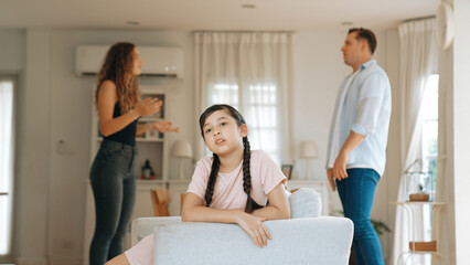 Annoyed and unhappy young girl sitting on sofa trapped in middle of tension by her parent argument...