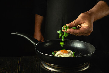 The cook adds chopped green onions to the pan with the eggs by hand. Concept of preparing breakfast with egg in restaurant kitchen