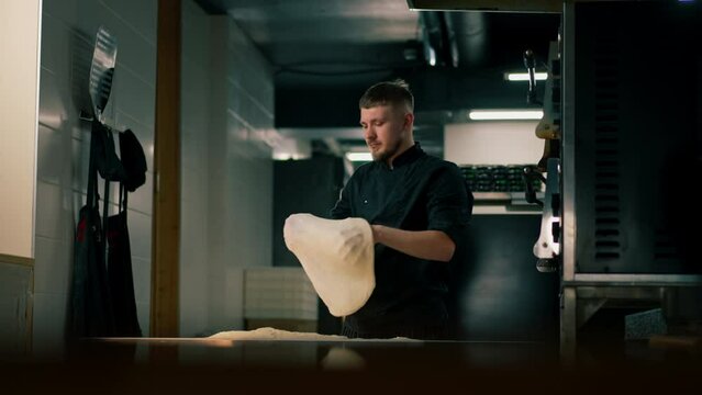 in a professional kitchen in a pizzeria young chef in a black uniform throws a pancake of dough into the air