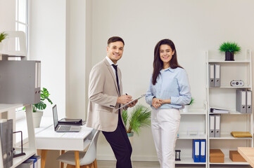 Portrait of two successful smiling business people man and woman standing in office near workplace and looking cheerful at camera. Coworkers and company employees working indoors together.