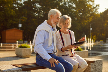 Portrait of happy smiling senior couple sitting outdoors on bench in city park using digital tablet for social media, searching information, watching funny movie or having online video call.