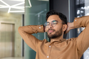 Close-up photo of a tired young Indian man resting in the office, taking a break from work, satisfied with the result, closed his eyes and threw his hands behind his head