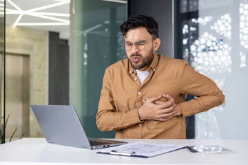 A young Indian man in a brown shirt is sitting at the desk in the office and is bent over from pain in the heart, holding his hands to his chest, feeling severe pain