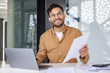 Portrait of a smiling young Muslim man working in the office with a laptop and documents, looking...