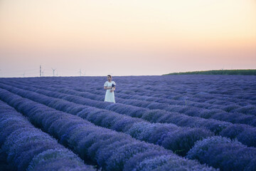 Woman in a white dress, walking among blooming lavender field in the golden hour at sunset.