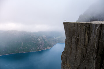 A lone figure stands on the edge of Preikestolen, a dramatic cliff in Norway, overlooking a vast expanse of water shrouded in mist. Preikestolen, Norway - Powered by Adobe