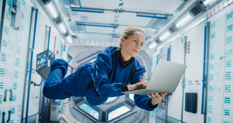 Female Astronaut Using Laptop Computer in Space Inside a Spacecraft. Working Online, Checking and Updating Flight Information, and Writing Report. Spacewoman Floating in Zero Gravity Inside Spaceship