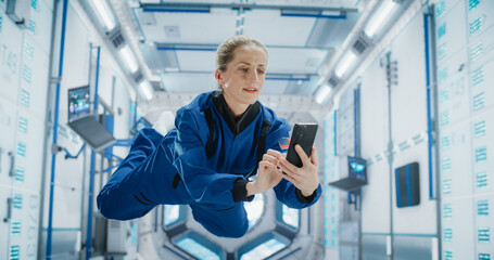 Female Astronaut Floating in Zero Gravity Inside a Space Station. Spacewoman Using a Smartphone in Space in Orbiting Spacecraft, Browsing Internet Content, Checking and Updating Social Media Networks