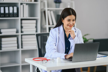 Asian doctor in a white lab coat is sitting at a desk with a laptop. She is smiling and she is happy