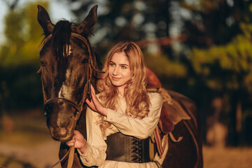 Obraz premium A young woman with a horse stands near a fence in autumn at sunset. Breeding thoroughbred horses for equestrian sport.