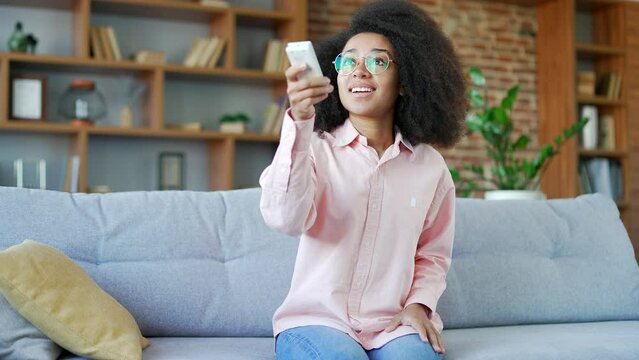 Happy young african american female turns on air conditioner by remote control sitting on sofa in living room at home. Smiling black woman presses button of device and turns off heating in the house