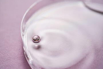 Serum or cosmetic oil flows into a transparent bowl on a purple background.