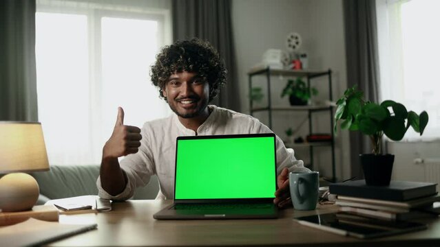 Smiling young indian man showing thumbs up sitting next to laptop computer with green chroma key screen advertising for your phone at home workplace Advise Approve Recommend Choice concept