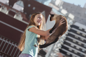 cheerful preteen girl lifting her cat up outdoors with city buildings in background