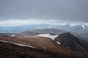 Dark overcast landscape with high pass, wide glacier on stony hill slope and snowy mountain range silhouette in far away in rainy low clouds. Large mountains with snow under gray sky in bad weather.