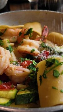 Putting grated parmesan cheese on a plate of paccheri al Profumo di Mare. The dish features al dente pasta, succulent shrimp, and a medley of fresh vegetables, all garnished with aromatic herbs for an
