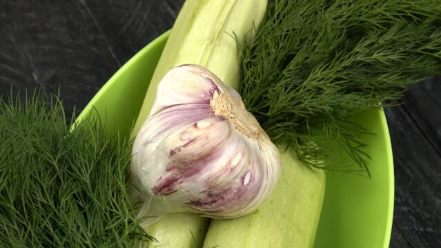 Fresh zucchini, bunch of dill and a head of young garlic in a green bowl, standing on a black wooden board