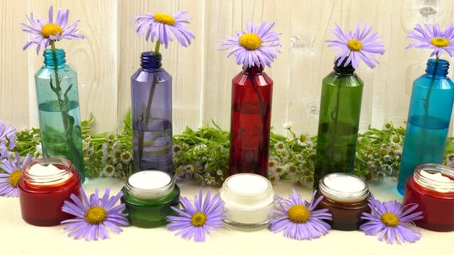 Purple asters like the chamomile placed in the colorful glass bottles with water, standing in a row. Jars with chamomile-based cosmetics stand on a white table against a background of fresh chamomile 