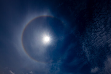 A captivating image of a sun halo against a deep blue sky, created by the refraction of sunlight through ice crystals in the atmosphere. The sun is bright, surrounded by a circular rainbow-like halo.