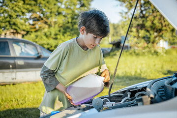 Fototapeta premium A young boy pours windshield washer fluid into a car's engine compartment on a sunny day. The scene is set outdoors with a blurred green background.