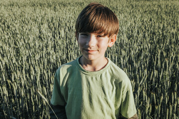 A young boy stands in the middle of a lush rye meadow, wearing a green shirt and smiling softly at the camera. The field stretches out behind him, creating a serene and natural backdrop.