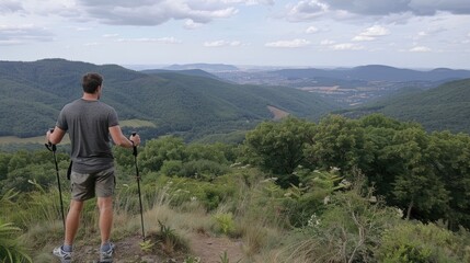 Naklejka premium A man with hiking sticks stands at the top of a mountain and looks at a forest landscape, mountains and sky in the distance.
