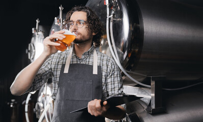 Fototapeta premium Expert brewer with clipboard in apron holds glass of craft beer and checking quality and color. Worker sommeliers taste drink on Brewery factory
