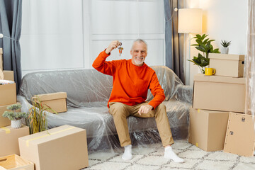 Smiling senior man holding keys from new home sitting sofa at cupboard boxes interior