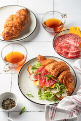 Croissant sandwich with salami sausage, cheese and cucumber in a plate with cup of tea for tasty breakfast on white tile table