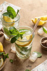 Two glasses of mojito cocktail or tonic with lemon, mint and ice on brown textured background. Refreshing summer drink