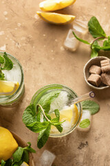 Two glasses of mojito cocktail or tonic with lemon, mint and ice on brown textured background. Refreshing summer drink, top view