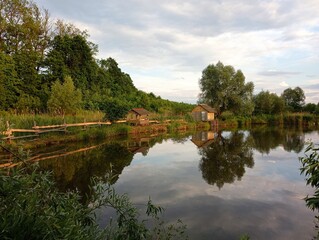 Obraz premium A cozy pond at sunset with wooden houses for relaxation and spending time in nature. A pond with trees on its banks and the reflection of a hut and trees on the surface of the water.