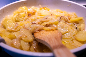 A plate of fresh potatoes and onion is fried in a white granite frying pan. wooden spoon for a bio, ecological, natural, healthy and delicious dish. It is the first part for a potato omelette receip