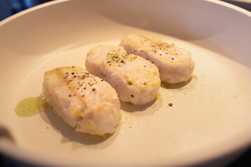 Three fresh hake loins are cooked in a white granite pan with extra virgin olive oil and aromatic herbs.