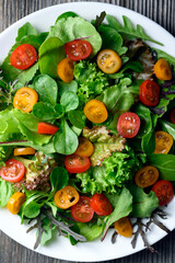 Mix of fresh organic juicy salad greens and red and yellow cherry tomato in white plate on rustic wooden table. Food photography