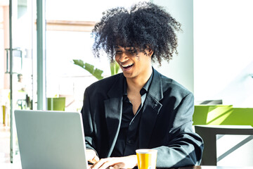 A non-binary person with an afro hairstyle, wearing a black suit, working with a white laptop and a yellow coffee cup while smiling