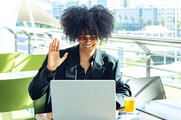 Non-binary person with afro hair, wearing a black suit, waving, looking at a laptop while smiling